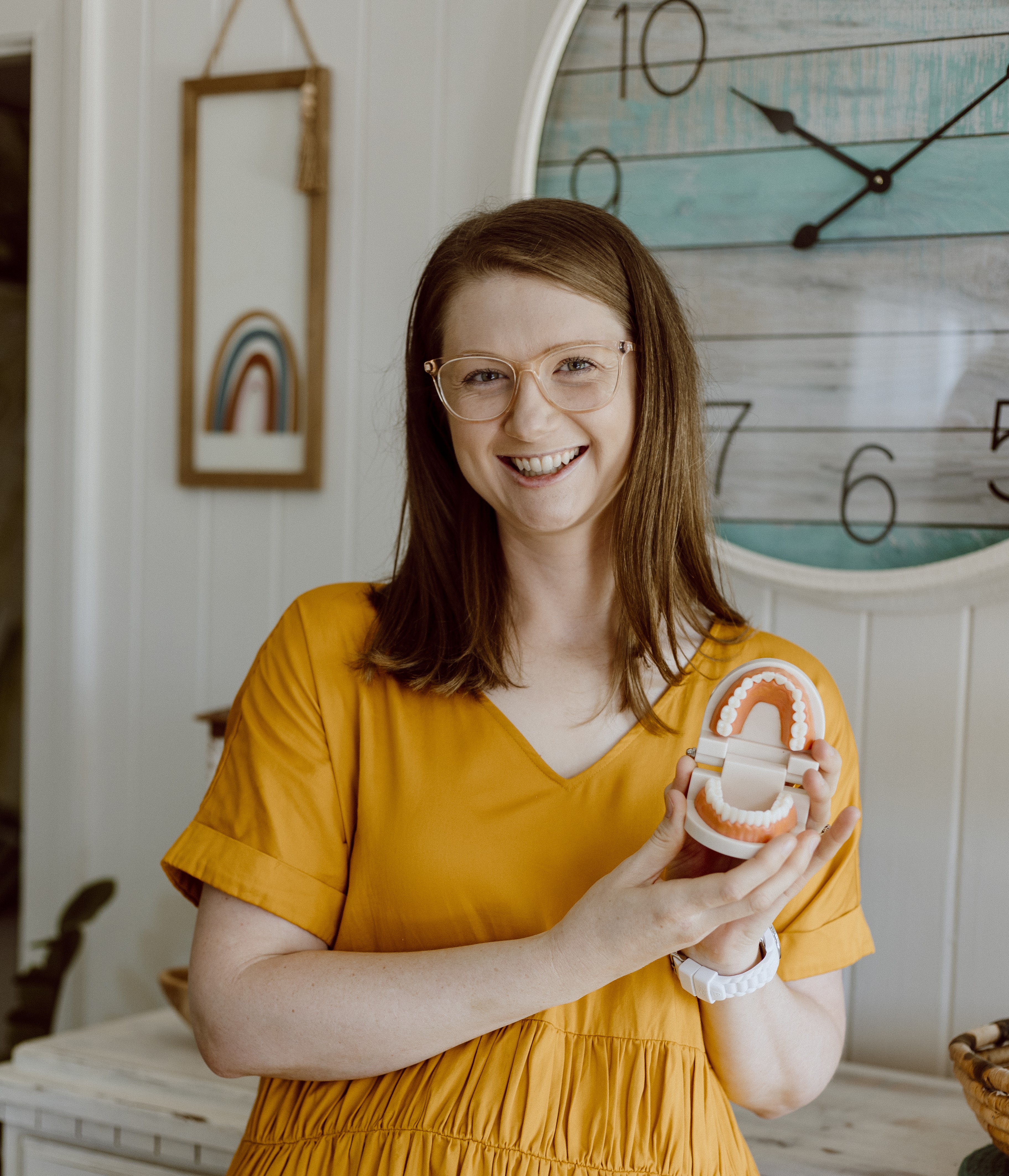 Mel Van Schelven, Oral Health Therapist and Orofacial Myofunctional Therapist at The Face Place, smiling and holding a dental model during a children's online oral health consultation.
