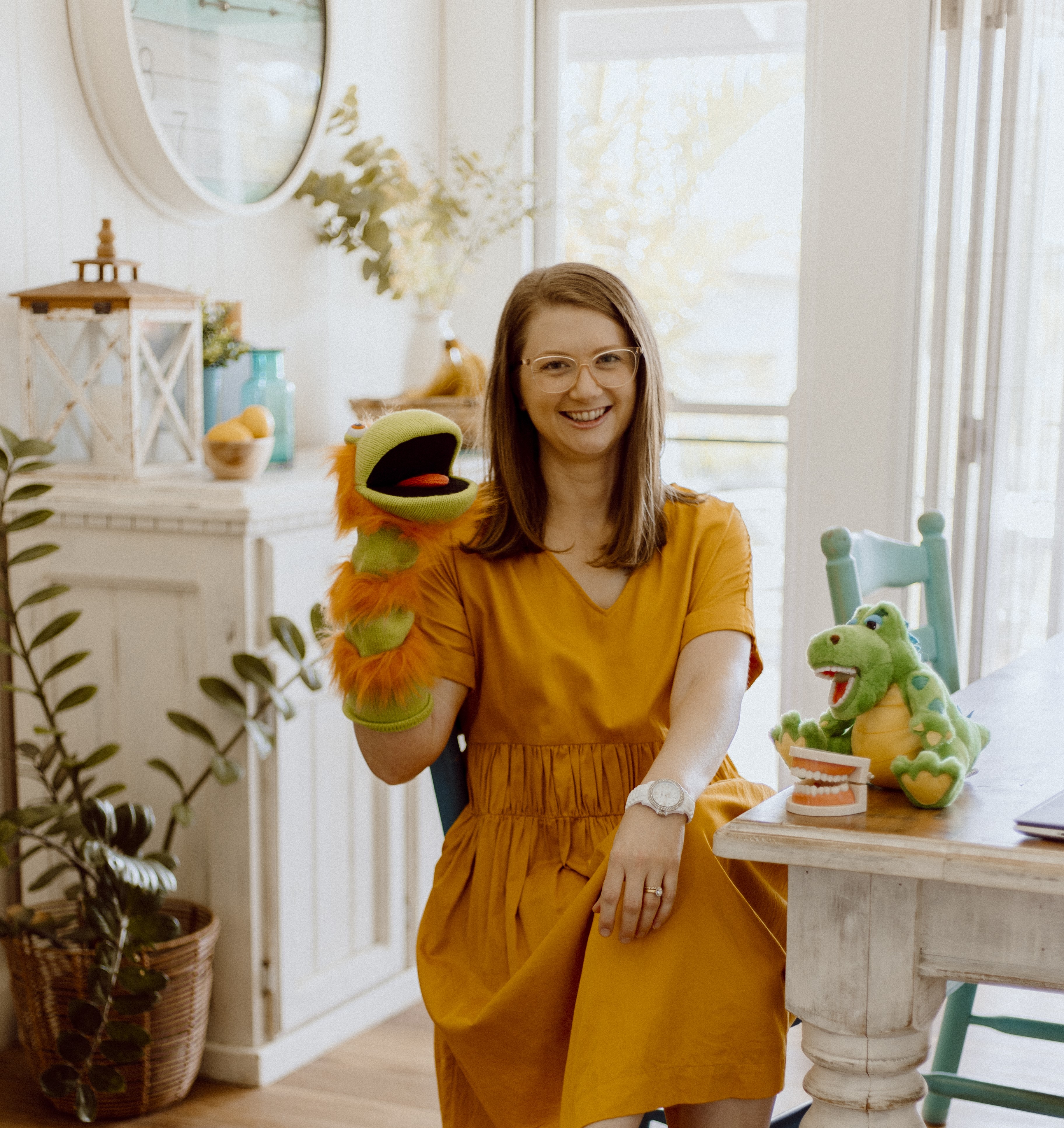 Oral health therapist Mel Van Schelven from The Face Place sitting in a bright home clinic, smiling with a puppet and children’s dental teaching toys, supporting families with children’s oral health and function.
